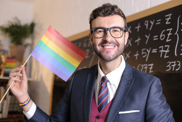 close-up portrait of handsome young teacher holding lgbtq flag in front of blackboard in classroom - Photo, Image
