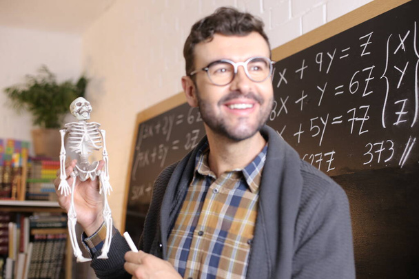 zbliżenie portret przystojny młody nauczyciel trzymając miniaturowy szkielet przed tablicą w klasie - Zdjęcie, obraz