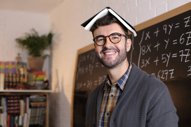 Nahaufnahme Porträt eines lustigen jungen Lehrers mit Buch auf dem Kopf vor der Tafel im Klassenzimmer - Foto, Bild