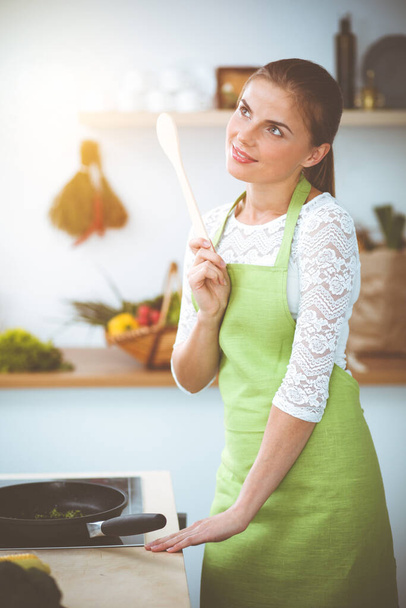 An attractive young dark-haired woman frying meal at worm pan by new keto recipe while standing and smiling in sunny kitchen. Cooking and householding concepts. - Fotoğraf, Görsel