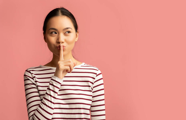 Guarda silencio. Joven mujer asiática sosteniendo dedo cerca de labios y mirando a un lado - Foto, Imagen