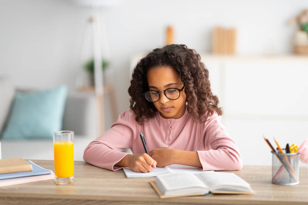 Nettes afrikanisch-amerikanisches Schulmädchen mit Brille, das in Notizblock schreibt, Hausaufgaben macht oder Essay schreibt, zu Hause lernt - Foto, Bild