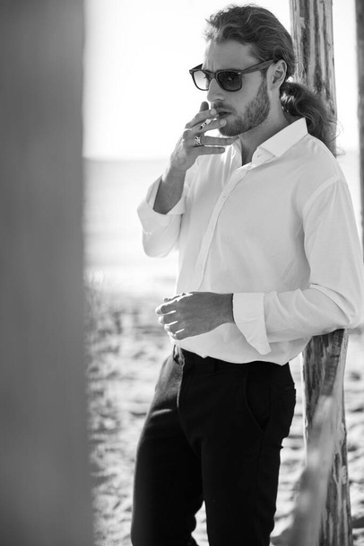 musta ja valkoinen kuva tyylikäs kaveri seisoo täydessä kasvussa ja valaistus savukkeen meren rannalla - Valokuva, kuva