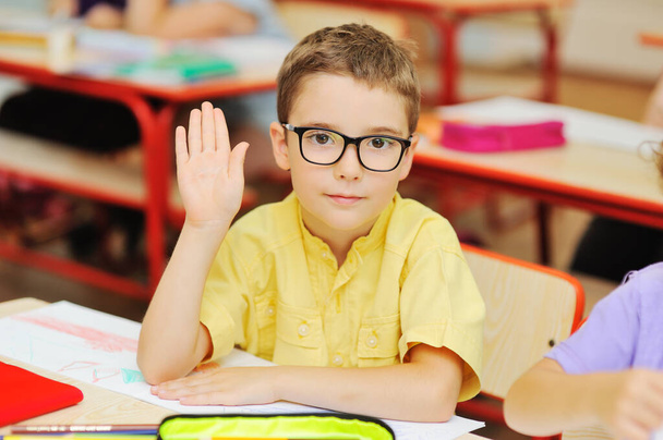 small child schoolboy in a yellow shirt and glasses reaches out for a response in the background of a primary school classroom. Back to school. - Photo, Image