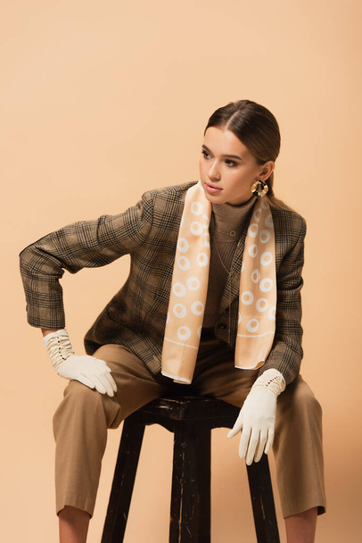 fashionable woman in blazer, pants and gloves looking away while sitting on stool isolated on beige - Photo, Image