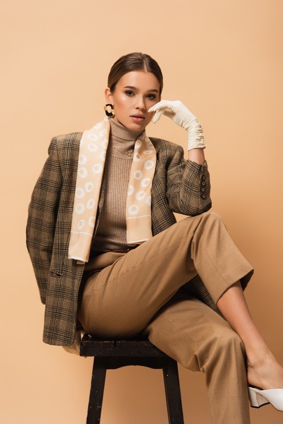 young woman in fashionable blazer and pants looking at camera while sitting on beige  - Foto, Imagem