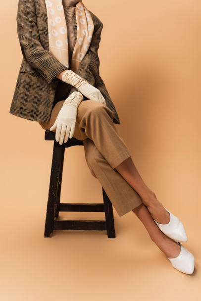 ořezaný pohled na ženu v kalhotách, kostkovaném blejzru, bílých rukavicích a botách sedících na béžové  - Fotografie, Obrázek