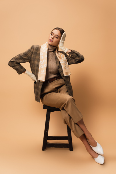 fashionable woman in brown blazer and trousers touching hair while sitting on beige  - Photo, Image