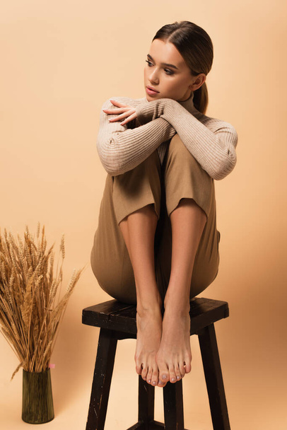 young woman in trendy pants and turtleneck sitting on chair barefoot on beige background - Photo, Image
