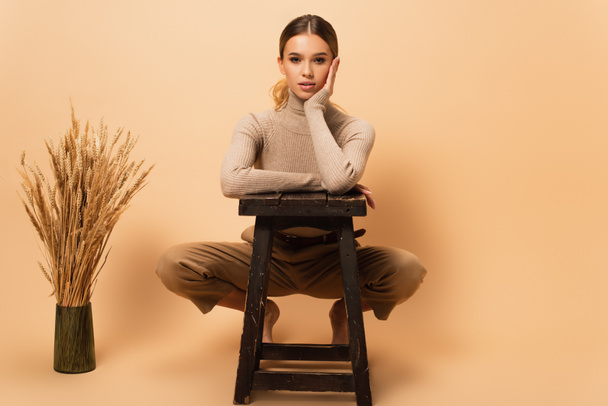 fashionable barefoot woman posing near stool and vase with spikelets on beige background - Foto, afbeelding