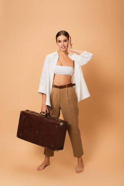barefoot, stylish woman with retro suitcase touching face while standing on beige background - Фото, изображение