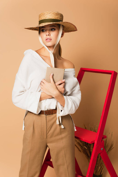 young woman in white shirt, trousers and straw hat holding book on beige background - Photo, Image