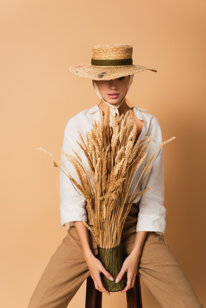 sensual woman in straw hat and white shirt holding vase of wheat spikelets and looking at camera on beige - 写真・画像