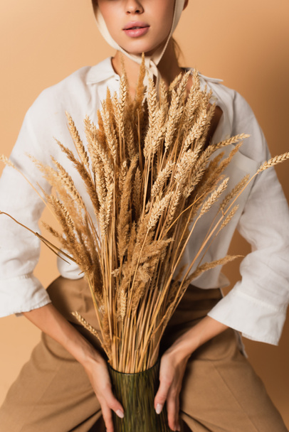 cropped view of young woman in white shirt holding wheat spikelets isolated on beige - Фото, зображення