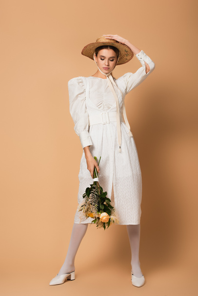 full length of young woman in dress and straw hat holding bouquet of flowers on beige - Foto, Imagem