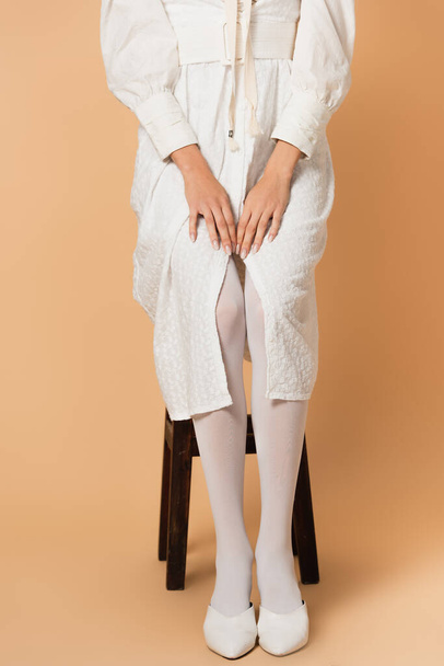 cropped view of young woman in white dress sitting on stool on beige - Foto, imagen