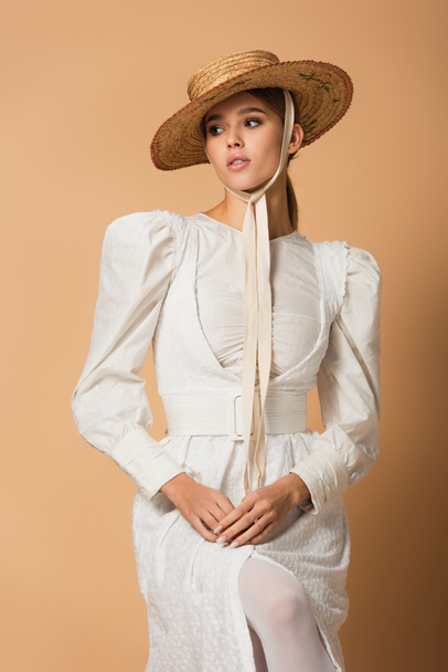 young woman in white dress and straw hat posing with clenched hands on beige - Foto, imagen
