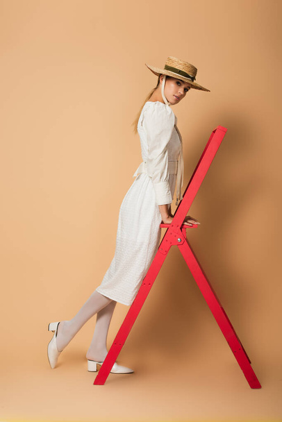 full length of young woman in white dress and straw hat standing on red ladder on beige - Photo, Image