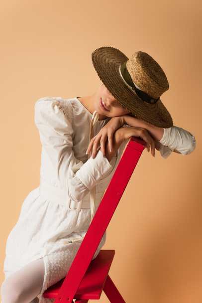 młoda kobieta w białej sukience i słomkowym kapeluszu opierająca się na czerwonej drabinie na beżu - Zdjęcie, obraz