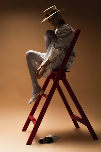 young woman in white dress and straw hat posing on red ladder in dark on beige - Foto, Imagem