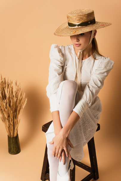 młoda kobieta w słomkowym kapeluszu pozuje na drewnianym krześle w pobliżu kolców pszenicy na beżu - Zdjęcie, obraz