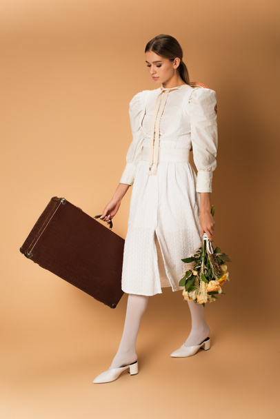 full length of young woman in white dress holding bouquet with flowers and suitcase on beige - Photo, Image
