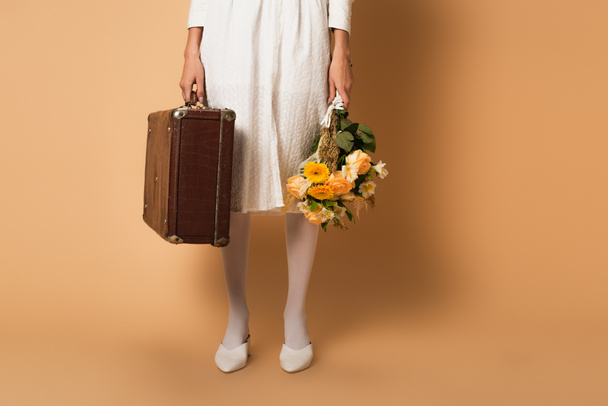 cropped view of young woman in white dress holding bouquet of flowers and suitcase on beige - Foto, imagen