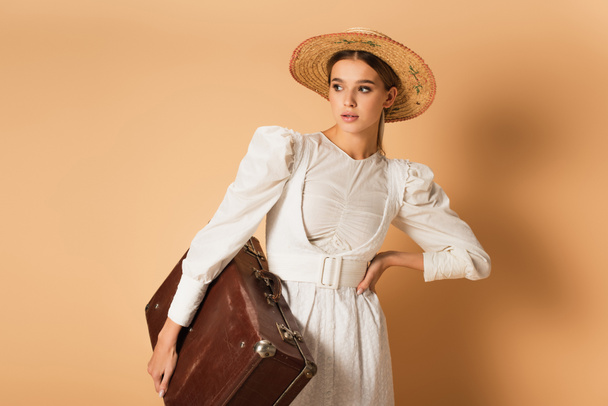 fiatal nő fehér ruhában és szalma kalap gazdaság bőrönd és pózol kézzel csípő bézs - Fotó, kép