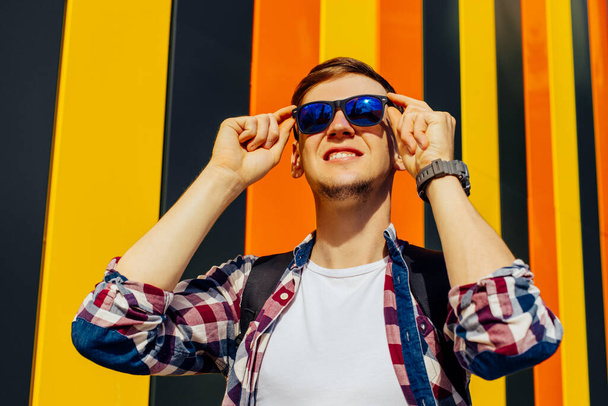 Happy young man, putting on sunglasses, in sunny weather, standing against the background of a colorful wall in the city - Zdjęcie, obraz