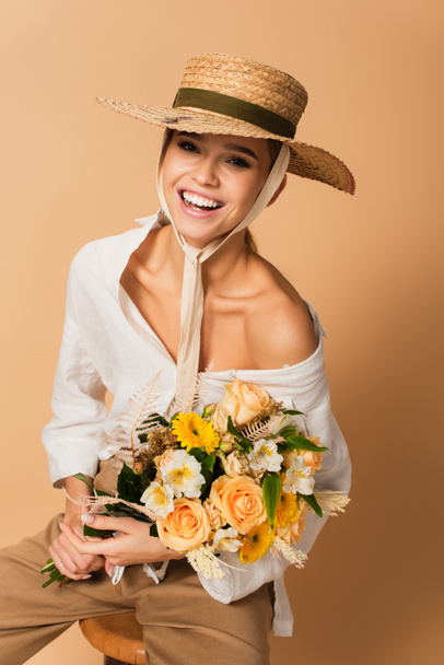 joyful young woman in straw hat holding bouquet of different flowers on beige - Fotoğraf, Görsel