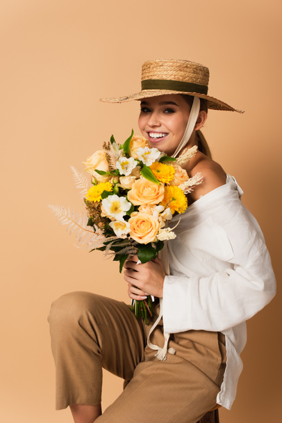 smiling young woman in white shirt and straw hat holding bouquet of flowers on beige - Photo, Image
