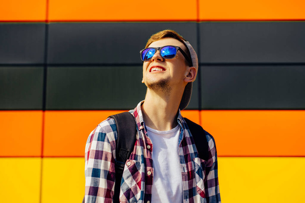Young happy smiling guy in sunglasses and a cap looking to the side posing while standing against a colored background of the city wall, People and Style concept - Fotó, kép