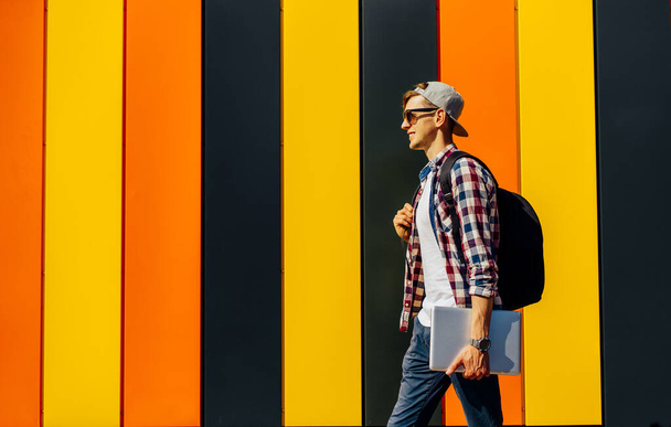 Stijlvolle jongeman met een aktetas, in een zonnebril en een pet, loopt langs een straat met een laptop, tegen de achtergrond van een kleurrijke muur, het concept van succes, het bedrijfsleven - Foto, afbeelding