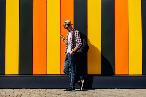 Young smiling stylish man in sunglasses and a cap, uses a mobile phone, leaning against a colorful wall, in the city - Fotografie, Obrázek