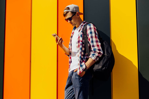 Young smiling stylish man in sunglasses and a cap, uses a mobile phone, leaning against a colorful wall, in the city - Foto, immagini