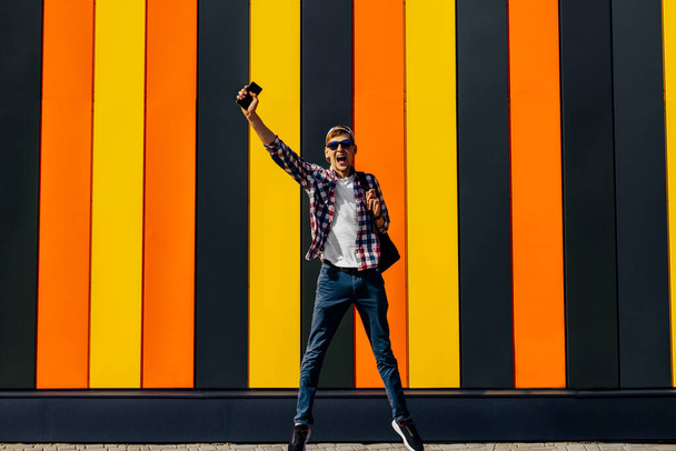 Excited happy young man, in sunglasses and a cap, shows a gesture of success, celebrating victory jumping up with a mobile phone, against the background of a bright colorful wall, on a city street - Foto, Bild