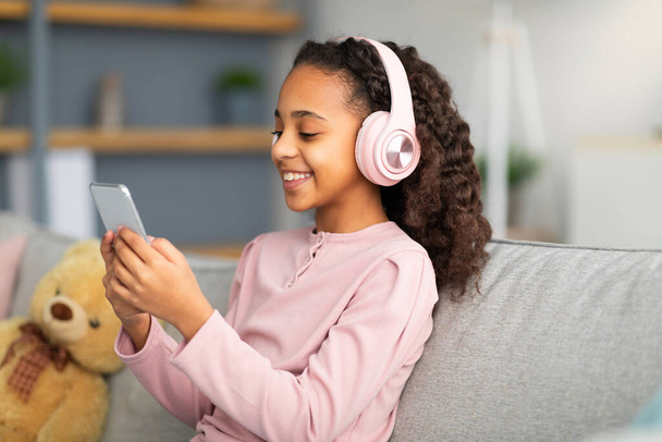 Музыкальное приложение. Радостная африканская американка слушает любимые песни в беспроводных наушниках, сидя дома на диване - Фото, изображение