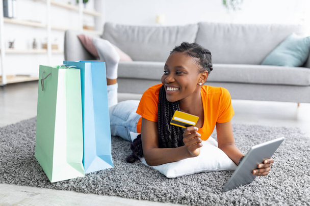 Shopaholism e e-commerce. Senhora preta com cartão de crédito compras on-line via laptop, deitado no chão com sacos dentro de casa - Foto, Imagem