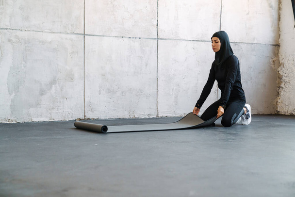 Young muslim woman in hijab unrolling fitness mat indoors - 写真・画像