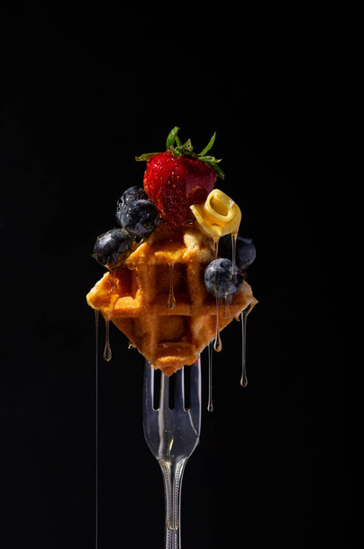 Belgian waffle with strawberry, blueberries and butter on a fork, topped with dropping honey. - Valokuva, kuva