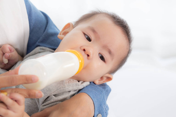 Jeune mère asiatique embrassant et nourrissant petite fille avec bouteille de lait à la maison, innocence du nouveau-né boire avec maman satisfaite, relation et liaison de maman et d'enfant, concept de famille. - Photo, image
