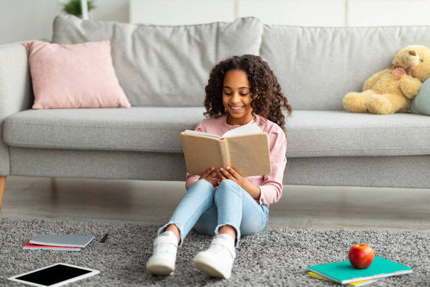Youth and literature. Smiling african american gen z girl holding and reading paper book, sitting on the floor carpet - Foto, Imagem