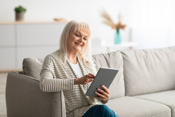 Улыбающаяся зрелая женщина с помощью планшета сидит на удобном диване - Фото, изображение