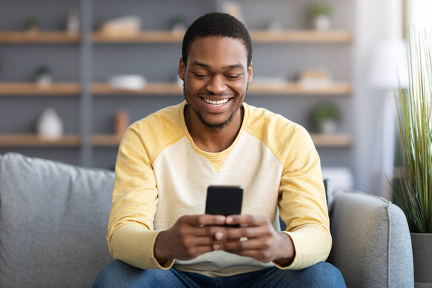 Smiling black guy sitting on couch, using smartphone - Фото, зображення