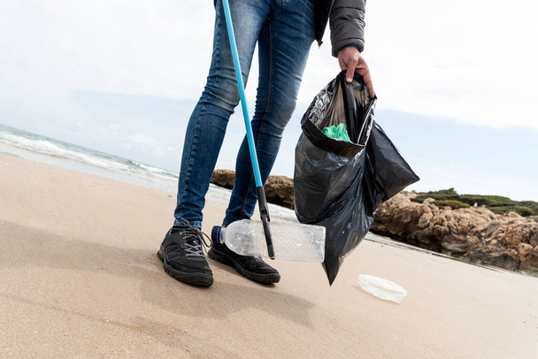 ένας άνθρωπος συλλέγει, με ένα εκτεινόμενο μέτρο, κάποια απόβλητα, όπως κονσέρβες, μπουκάλια ή σακούλες, από την άμμο μιας μοναχικής παραλίας, ως δράση για τον καθαρισμό του φυσικού περιβάλλοντος - Φωτογραφία, εικόνα