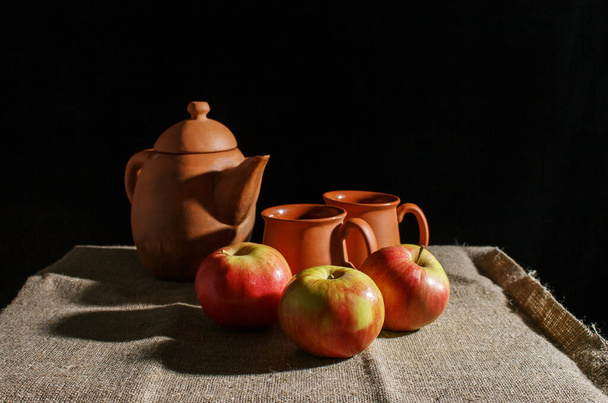 Martwa natura z glinianym czajnikiem, dwoma filiżankami i trzema czerwonymi jabłkami. wewnętrzne zbliżenie na czarnym tle - Zdjęcie, obraz