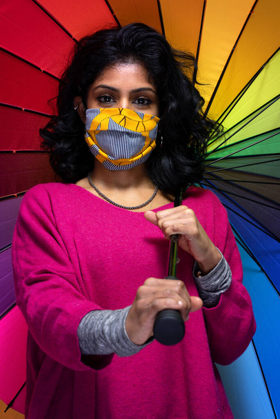 Belle jeune femme indienne posant avec un parapluie coloré avec une protection de masque covid 19 - Jolie fille Sri Lanka utilise la protection contre la grippe pandémique tout en se couvrant d'un grand parapluie couleur - Photo, image