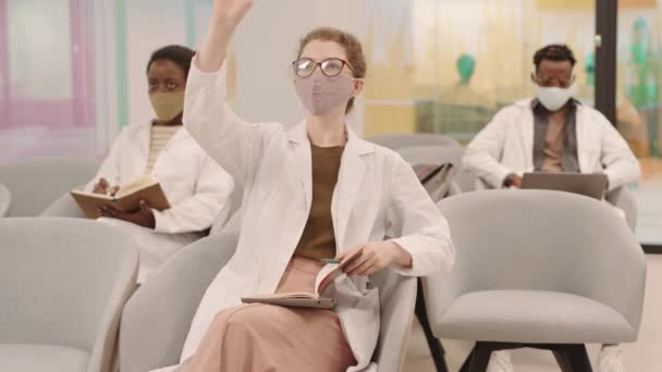 Середня повільність трьох молодих багатоетнічних студентів-медиків у білих халатах та масках для обличчя, що роблять нотатки в книзі або ноутбуці, слухаючи лекції в сучасному класі Дівчина піднімає руку, щоб поставити запитання
 - Кадри, відео
