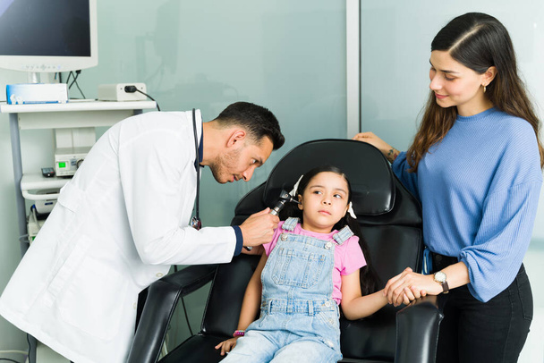 I don't like coming to the doctor. Scared little girl sitting at the examination chair and getting checked due to an ear infection - 写真・画像
