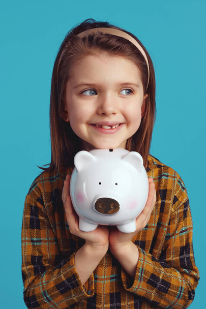 Χαριτωμένο χαρούμενο παιδί κρατώντας το κουτί με τα χρήματα σε σχήμα γουρουνιού κοντά στο πρόσωπο κοιτάζοντας στην άκρη - Φωτογραφία, εικόνα